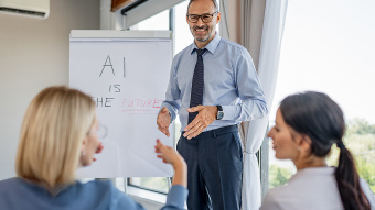 Ein Mann im Business-Outfit steht vor einem Flipchart in einem hellen Besprechungsraum und spricht zu zwei sitzenden Kolleginnen. Auf dem Flipchart steht der handschriftliche Text „AI is the future“. Der Mann gestikuliert während der Präsentation, die beiden Frauen hören aufmerksam zu und beteiligen sich am Gespräch. Im Hintergrund sind große Fenster mit Tageslicht zu sehen, die eine moderne Arbeitsumgebung vermitteln.