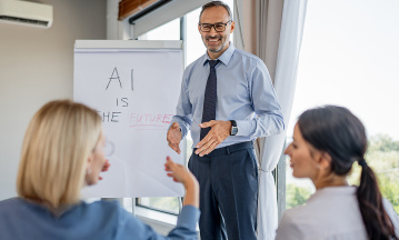Ein Mann im Business-Outfit steht vor einem Flipchart in einem hellen Besprechungsraum und spricht zu zwei sitzenden Kolleginnen. Auf dem Flipchart steht der handschriftliche Text „AI is the future“. Der Mann gestikuliert während der Präsentation, die beiden Frauen hören aufmerksam zu und beteiligen sich am Gespräch. Im Hintergrund sind große Fenster mit Tageslicht zu sehen, die eine moderne Arbeitsumgebung vermitteln.