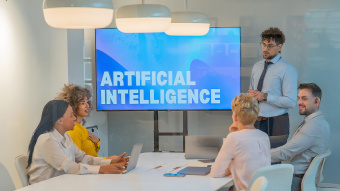 Vier Menschen sitzen um einen Konferenztisch, eine fünfte männliche Person steht hinter dem Konferenztisch neben einem Screen, auf dem mit weißer Schrift "Artificial Intelligence" steht. 