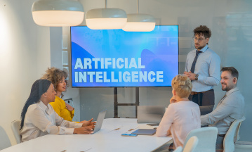Vier Menschen sitzen um einen Konferenztisch, eine fünfte männliche Person steht hinter dem Konferenztisch neben einem Screen, auf dem mit weißer Schrift "Artificial Intelligence" steht. 