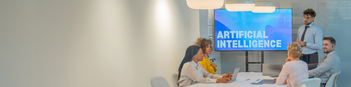 Vier Menschen sitzen um einen Konferenztisch, eine fünfte männliche Person steht hinter dem Konferenztisch neben einem Screen, auf dem mit weißer Schrift "Artificial Intelligence" steht. 
