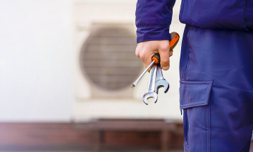 Servicetechniker in Arbeitskleidung hält Werkzeug in der Hand und steht vor einer Wärmepumpe.