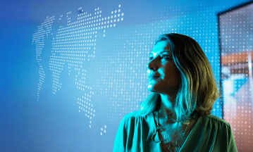 Frau im Profil blickt nach rechts vor einem blauen, digitalen Hintergrund mit abstrahierter Weltkarte aus Punkten. Die Szene vermittelt Zukunftsorientierung, Datenanalyse und technologische Transformation im Audit-Umfeld.