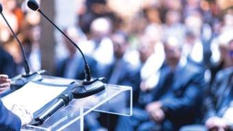 DAs Bild zeigt Menschen, die auf Stühlen sitzen und offensichtlich einem Speaker zuhören, denn im Vordergrund sind Mikrofone zu sehen. 