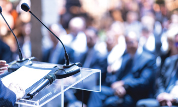 DAs Bild zeigt Menschen, die auf Stühlen sitzen und offensichtlich einem Speaker zuhören, denn im Vordergrund sind Mikrofone zu sehen. 