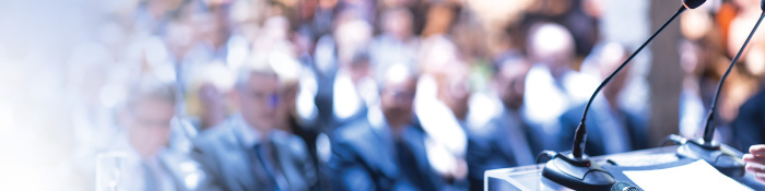 DAs Bild zeigt Menschen, die auf Stühlen sitzen und offensichtlich einem Speaker zuhören, denn im Vordergrund sind Mikrofone zu sehen. 