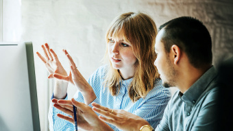 Eine Frau und ein Mann stehen vor einem Computermonitor und richten ihre offenen Handflächen zum Bildschirm.