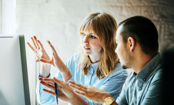 Eine Frau und ein Mann stehen vor einem Computermonitor und richten ihre offenen Handflächen zum Bildschirm.