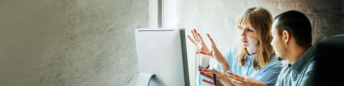 Eine Frau und ein Mann stehen vor einem Computermonitor und richten ihre offenen Handflächen zum Bildschirm.