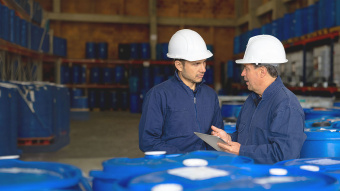 Zwei Mitarbeiter mit Schutzhelmen stehen in einer Lagerhalle der Chemieindustrie zwischen großen blauen Fässern. Sie betrachten gemeinsam ein Tablet und besprechen offenbar Produktions- oder Sicherheitsdaten, während im Hintergrund weitere Fässer in Regalen gelagert sind.