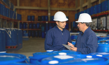 Zwei Mitarbeiter mit Schutzhelmen stehen in einer Lagerhalle der Chemieindustrie zwischen großen blauen Fässern. Sie betrachten gemeinsam ein Tablet und besprechen offenbar Produktions- oder Sicherheitsdaten, während im Hintergrund weitere Fässer in Regalen gelagert sind.