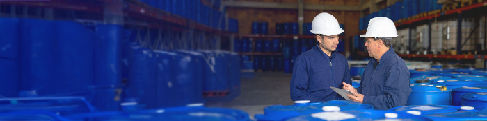 Zwei Mitarbeiter mit Schutzhelmen stehen in einer Lagerhalle der Chemieindustrie zwischen großen blauen Fässern. Sie betrachten gemeinsam ein Tablet und besprechen offenbar Produktions- oder Sicherheitsdaten, während im Hintergrund weitere Fässer in Regalen gelagert sind.