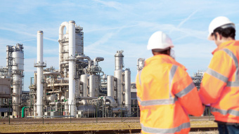 Zwei Fachkräfte in Schutzkleidung stehen vor einer großflächigen Chemieanlage mit Rohrleitungen, Türmen und Industrieanlagen unter blauem Himmel.