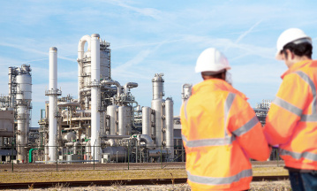 Zwei Fachkräfte in Schutzkleidung stehen vor einer großflächigen Chemieanlage mit Rohrleitungen, Türmen und Industrieanlagen unter blauem Himmel.