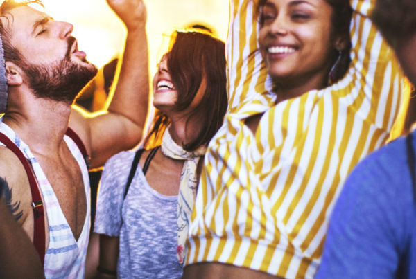 Closeup of group of multi ethnic young adults dancing and enjoying an open air concert on a summer night. There are three girls and three guys in a candid shot against bright background light.