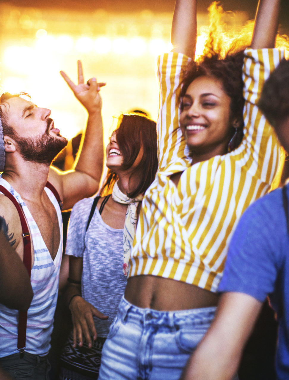 Closeup of group of multi ethnic young adults dancing and enjoying an open air concert on a summer night. There are three girls and three guys in a candid shot against bright background light.